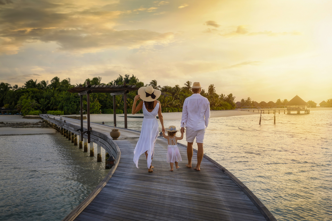 A family walks holding hands over a wooden pier towards a tropical island